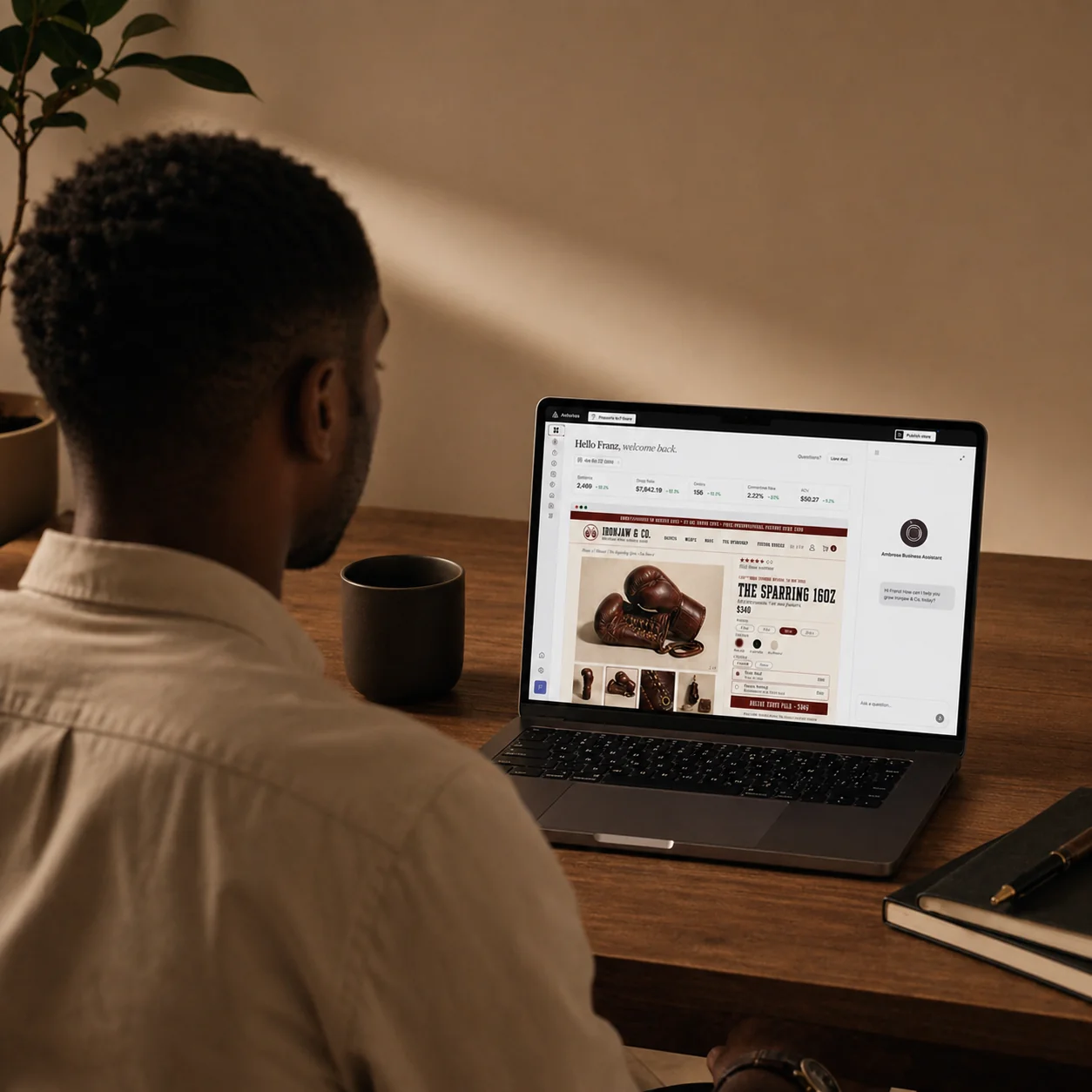 A merchant at a MacBook reviewing the Amboras admin: real-time KPIs, an embedded Ironjaw & Co storefront preview, and the Amboras Business Assistant chat docked on the right
